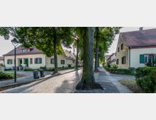 Straßenszene in einem Wohngebiet mit gelb verputzten Häusern im traditionellen Baustil, roten Ziegeldächern und grünen Fensterläden. Bäume säumen die gepflasterte Straße, die von einem Gehweg und niedrigen Hecken begleitet wird.
