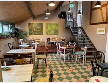 Gemütliches Café mit Holzmöbeln und buntem Stuhlmix auf einem grünen und beigen Schachbrettmusterboden. Im Hintergrund sind Pflanzen, zwei großformatige Malereien und eine Treppe aus Metall zu sehen.