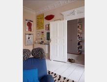 Eclectic apartment living room with white-painted floors, ornate ceiling molding, and a tall white paneled door opening into an adjoining room. The walls are covered with framed art and a red lips wall sculpture, with blue seating, patterned cushions, and shelving visible through the doorway.