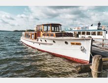Ein weißes Holzboot mit braunen Akzentelementen und dem Namen "Don Juan" liegt am Ufer eines Sees, festgemacht an einem Holzsteg. Im Hintergrund ist ein größeres Passagierschiff sowie eine bewaldete Uferlandschaft zu sehen, unter einem Himmel mit vielen Wolken.