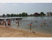 Sandstrand mit zahlreichen Badegästen im flachen Wasser eines Sees. Im Hintergrund sind Bäume, ein Steg, moderne Gebäude und ein markantes rotes Gebäude am Ufer zu sehen.