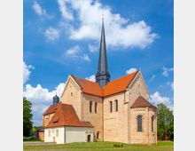 Eine Backsteinkirche mit einem hohen, schlanken Kirchturm und roten Ziegeldächern, umgeben von einer Wiese unter einem blauen Himmel mit weißen Wolken. Die Architektur weist Rundbogenfenster und romanische Stilelemente auf.
