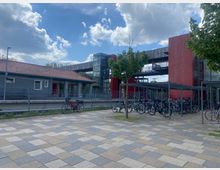 Bahnhofsumgebung mit Fahrradabstellplatz, gekennzeichnet durch ein modernes Gebäude mit roten Fassaden, einer Glasfassade mit Aufzug und einem überdachten Bereich für Fahrräder. Im Vordergrund ist ein gepflasterter Platz zu sehen.