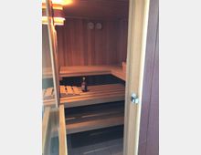 Holzverkleideter Saunaraum mit zwei Sitzbänken in L-Form, einer Glas-/Holztür, einer Flasche auf der oberen Bank und zwei Wandthermometern zur Temperaturanzeige. Der Boden ist gefliest.