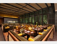 Elegantes Restaurant mit gepolsterten Stühlen und sofas in warmen Gelb- und Grüntönen sowie dunklen Holztischen, umgeben von bodentiefen Fenstern mit Blick auf eine beleuchtete grüne Pflanzenwand. Eine offene Küche befindet sich im Hintergrund, abgetrennt durch eine graue Steinwand.