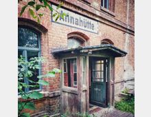 Backsteingebäude mit der Aufschrift "Annahütte" über einem alten hölzernen Eingangsbereich, flankiert von Fenstern mit Rundbögen; umgeben von Pflanzenbewuchs.