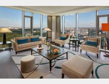 Wohnzimmer in einem modernen Hochhaus mit großer Fensterfront, die eine Panoramaansicht über eine Stadt und einen Park bietet. Die Einrichtung umfasst beige Polstermöbel, blaue Kissen, einen Teppich mit dezentem Muster, Tischlampen und einen Couchtisch mit Dekorationen wie Blumen und Früchten.