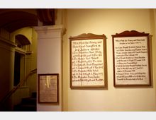 Innenraum einer historischen Kirche oder Gedenkstätte mit einer cremefarbenen Wand, an der gerahmte Tafeln mit handgeschriebenem Text in deutscher Schrift angebracht sind. Im Hintergrund befindet sich ein Treppenaufgang mit Rundbogenarchitektur und dezentem Lichteinfall.