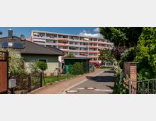 Wohngebiet mit einem einstöckigen Einfamilienhaus im Vordergrund, das ein dunkles Ziegeldach und eine grüne Fassade hat. Im Hintergrund steht ein mehrstöckiges Wohngebäude mit Balkonen, umgeben von Gärten, Bäumen und einem gepflasterten Weg.