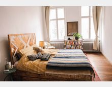 Schlafzimmer mit Holzfußboden, einem Bett mit dekorativem Holzkopfteil, bunten Kissen und Tagesdecke. Im Hintergrund ein Bücherregal, ein Sessel, ein Beistelltisch mit Lampe und Fenster mit weißen Vorhängen, die Licht hineinlassen.