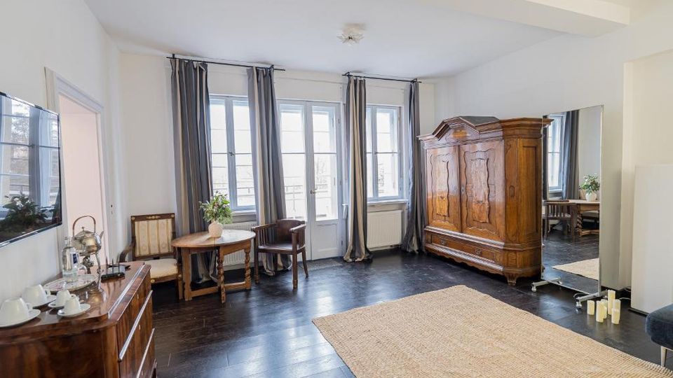 Heller Wohn- oder Hotelzimmerraum mit dunklem Holzboden, großen Fenstern und Balkontür mit langen Vorhängen. Im Raum stehen ein antiker Holzschrank, kleine Sitzgruppe mit Tisch, Kommode mit Tablett sowie ein großer Standspiegel neben einem Teppich.