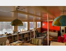 Innenraum eines Hausboots mit Blick auf das Wasser durch große rechteckige Fenster. Der Raum ist hell und enthält Möbel wie einen Esstisch mit Stühlen, Bücherregale, bunte Dekorationen und Lampen mit Globus-Motiv.