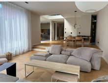 Modernes Wohnzimmer mit offener Raumgestaltung, hellen Wänden und großen bodentiefen Fenstern mit weißen Vorhängen. Im Vordergrund steht ein gemütliches Sofa, dahinter ein Essbereich mit Holztisch und Stühlen; im Hintergrund ist eine offene Küche sichtbar.