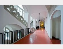 Weitläufiger Flur in einem historischen Gebäude mit roten Böden, weißen Wänden und hohen Gewölbedecken. Auf der linken Seite führt eine geschwungene Treppe nach oben, begleitet von kunstvoll verzierten Elementen, und der Gang ist mit schwarzen Geländern sowie Laternenleuchten dekoriert.