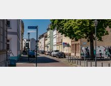 Stadtstraße mit parkenden Autos und mehrstöckigen Wohnhäusern, teils mit farbigen Fassaden. Rechts am Straßenrand stehen große Bäume sowie ein Gebäude mit graffitiverzierter Wand.