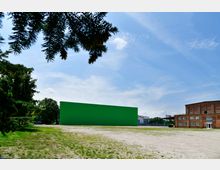 Freifläche mit Gras und Sand, im Hintergrund eine große, grüne Wand sowie ein Backsteingebäude mit mehreren Fenstern; am linken Bildrand sind Bäume und Tannenzweige zu sehen. Der Himmel ist blau mit einigen Wolken.