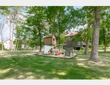 Ein Spielplatz mit einem Holzspielhäuschen, Kletterwand und Schaukel auf einer sandbedeckten Fläche, umgeben von hohen Bäumen und einer grünen Wiese. Im Hintergrund stehen zwei Wohnhäuser, eines davon dunkelgrau mit roten Dachziegeln.