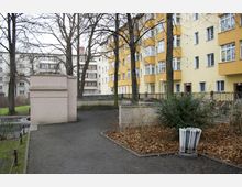 Wohnhof mit gelb gestrichenen Mehrfamilienhäusern, umgeben von kahlen Bäumen. Im Vordergrund ein gepflasterter Weg, ein Mülleimer sowie ein kleiner bepflanzter Bereich mit Sträuchern.