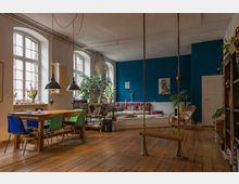 Wohnraum in einem industriellen Loft-Stil mit großen Sprossenfenstern, Holzboden und hohen Decken. Im Raum befinden sich ein Holztisch mit bunten Stühlen, eine Sitzecke mit Kissen vor einer blauen Akzentwand, zahlreiche Pflanzen sowie eine Holzschaukel an Seilen.
