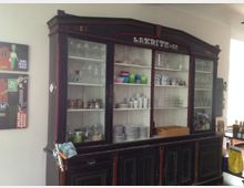 Ein antiker, dunkler Holzschrank mit Glastüren, gefüllt mit Geschirr, Gläsern und Vorratsgläsern, steht in einem hellen Raum. Links daneben hängen gerahmte Poster an der Wand.