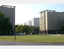 Platz mit einer Rasenfläche im Vordergrund, flankiert von zwei mehrstöckigen Gebäuden aus Stein mit regelmäßigen Fensterfassaden. Im Hintergrund sind Bäume und ein modernes, weißes Gebäude sichtbar, sowie parkende Autos und eine Straße.