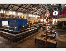 Ein großzügiger, moderner Gastronomiebereich in einer umgebauten Industriehalle mit einem auffälligen blauen Container als zentrale Bar. Der Raum ist geprägt von hohen Decken, industriellen Elementen, bunten Wandgraffiti und arrangierten Sitzbereichen mit Tischen und Stühlen.