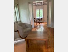 Wohnraum mit Holzboden und einfach eingerichteter Möblierung. Im Vordergrund stehen Sessel und ein Holztisch, und im Hintergrund führt eine Tür mit Glaseinsatz in einen weiteren Raum mit einem Tisch und Stühlen vor einem Fenster mit Blick ins Grüne.