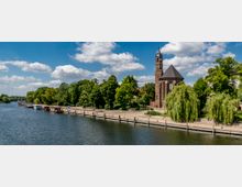 Uferpromenade an einem Fluss mit einer alten Backsteinkirche samt Glockenturm im Hintergrund, umgeben von Bäumen und grüner Vegetation. Der Bereich ist mit Weidenbäumen, Fußwegen und kleinen Bootsanlegern gestaltet, unter einem Himmel mit vereinzelten Wolken.