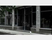 Außenansicht eines modernen Gebäudes mit großen Glasfenstern und einem Eingangsbereich aus Metall und Glas. Auf zwei Fenstern ist der Schriftzug „PLATTE“ zu sehen, und im Vordergrund befinden sich ein Gehweg und einige Baumzweige.