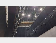 Blick auf eine Bühnendecke eines Theaters oder Studios mit zahlreichen Scheinwerfern und Beleuchtungsgeräten, die an Traversen befestigt sind. Im Hintergrund sind Metallgerüste und technische Installationen sichtbar.