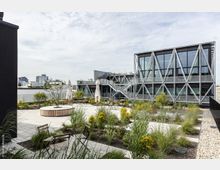 Moderne Dachterrasse mit bepflanzten Beeten, Sitzmöglichkeiten und Sonnenschirmen, umgeben von einem Gebäude mit Stahlträger-Fassade und großen Glasfenstern. Im Hintergrund sind weitere Gebäude und ein bewölkter Himmel sichtbar.