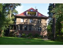 Zweistöckiges Wohnhaus mit Putzfassade und rotem Satteldach, umgeben von einem Garten mit Rasen, Blumenbeeten und hohen Bäumen. Auf einem Balkon im Dachgeschoss stehen Pflanzen, und eine Liege ist im Vordergrund aufgestellt.