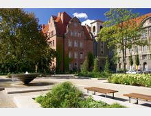 Eine parkähnliche Anlage mit einem modernen Brunnen im Vordergrund, umgeben von Bänken und grünen Bepflanzungen. Dahinter sind historische Gebäude aus rotem Backstein und grauem Stein mit verzierten Fassaden und roten Dächern zu sehen.