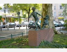 Bronzestatue eines Jungen, der einen Hund an einer Leine hält, auf einem roten Backsteinsockel vor einem großen Baum; im Hintergrund eine gepflasterte Straße, parkende Autos und Wohngebäude mit Balkonen.