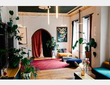 Wohnzimmer mit Holzboden und einer Mischung aus modernen und vintage Elementen. Der Raum enthält Pflanzen, Wandbilder, einen roten Teppich, bunte Sitzmöbel und einen Rundbogen mit roten Vorhängen.