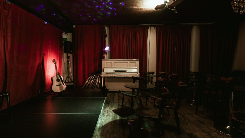 Small indoor music venue with a low stage in front of red curtains, featuring a white upright piano and an acoustic guitar near stage lighting. Wooden floors, scattered chairs and tables, and ceiling-mounted spotlights with colored light reflections define the intimate performance space.