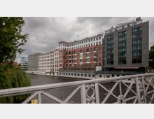 Mehrstöckige Gebäude mit moderner und klassischer Architektur, darunter rote Ziegel- und Glasfassaden, entlang eines Flusses. Im Vordergrund steht ein verzierter Metallzaun einer Brücke, umgeben von Bäumen und bewölktem Himmel.