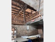 Innenansicht eines alten Gebäudes mit unverputzten Ziegelwänden und Holzbalkenkonstruktion. Sichtbare Zwischendecke aus Holz, Kabelstränge an Decke und Wänden sowie einfache Möbel und gestapelte Gegenstände im Raum.