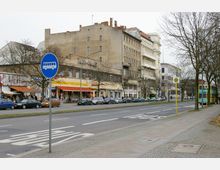 Straßenszene mit mehrspuriger Fahrbahn und Gehweg, gesäumt von älteren Wohn- und Geschäftshäusern. Im Vordergrund befindet sich ein Haltestellenschild sowie ein Bushaltesymbol auf der Straße; links sind parkende Autos und Geschäfte mit Werbeschildern erkennbar.