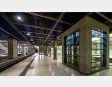 Moderne Bahnhofsplattform mit glatten, hellen Böden, säulenartigen Betonstützen und einem verglasten Aufzugsschacht auf der rechten Seite. Im Hintergrund sind Gleise, Sitzbereiche und blaue Hinweisschilder zu sehen, umgeben von metallischen Deckenstrukturen.