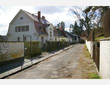 Wohnstraße mit alten Einfamilienhäusern, hell verputzt, roten Ziegeldächern und dekorativen Fenstern. Die Straße ist schmal und uneben, gesäumt von Zäunen und Hecken, mit Bäumen im Hintergrund.