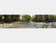 Straßenszene in einer urbanen Umgebung mit Kopfsteinpflaster, flankiert von Wohn- und Geschäftsgebäuden sowie Bäumen. Im Vordergrund stehen Fahrräder, Poller und ein roter Hydrant, während die Straße von geparkten Autos gesäumt ist.