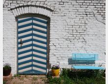 Weiße Ziegelsteinwand mit einer Holztür in Zickzack-Muster aus blau-weißen Streifen und einem antiken Türgriff. Daneben steht eine hellblaue Bank, flankiert von zwei Blumentöpfen mit violetten Blumen auf einem gepflasterten Boden.