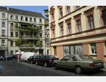 Straßenszene mit historischen Mehrfamilienhäusern im Altbaustil; das linke Gebäude hat Fassadendetails, Balkone mit Pflanzenbewuchs, während das rechte Gebäude sandgelb gestrichen ist. Auf der gepflasterten Straße parken Autos, darunter ein grüner Mercedes, und einige Passanten sind zu sehen.