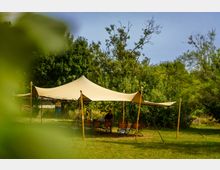 Weißes Sonnensegel auf einer Wiese mit Holztischen und -stühlen darunter, umgeben von dichtem Grün und Bäumen im Hintergrund.