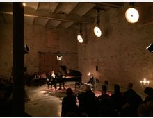 Ein kleiner Konzertsaal mit unverputzten Backsteinwänden, niedriger Decke und dimmer Beleuchtung. In der Mitte steht ein Flügel, umgeben von einem sitzenden Publikum, mit mehreren Kerzenleuchtern und punktueller Bühnenbeleuchtung.