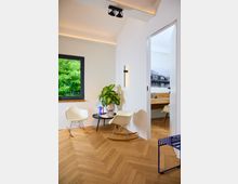 Heller Innenraum mit Parkettboden und Wänden in Weiß, eingerichtet mit zwei hellen Schaukelstühlen, einem kleinen runden Tisch mit einer Zimmerpflanze und Büchern. Ein Fenster mit Blick auf grüne Bäume und eine Türöffnung führen zu einem weiteren Raum mit minimalistischer Einrichtung.