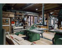 Eine Holzwerkstatt mit mehreren Maschinen, darunter eine grüne Tischkreissäge, sowie Regalen mit Werkzeugen und Holzmaterialien. Im Hintergrund sind Fenster mit Blick nach draußen und Holzstapel sichtbar, die Beleuchtung erfolgt durch Deckenlampen.