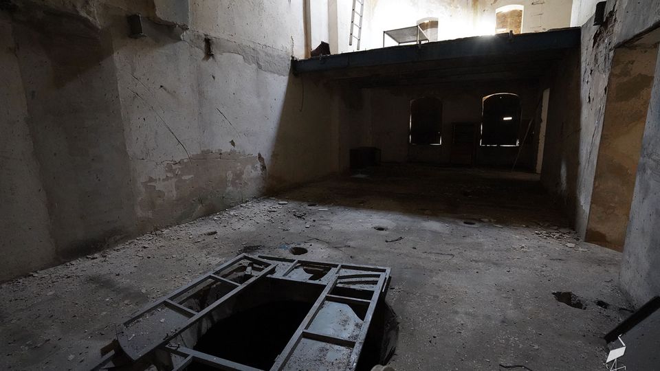 Innenraum eines verfallenen Industriegebäudes mit hohen, abblätternden Wänden, einer Zwischenebene und Licht, das durch Öffnungen im oberen Bereich einfällt. Auf dem staubigen Betonboden befindet sich eine große rechteckige Bodenöffnung mit Metallrahmen, im Hintergrund sind dunkle Türen oder Fensteröffnungen zu sehen.