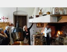 Eine Gruppe von Personen steht in einer rustikalen Küche mit einem offenen Kamin. Der Raum ist mit Holzmöbeln, Kupfergeschirr und Dekorationen wie einem Hirschdekor auf einem Regal gestaltet, während eine Frau gestikulierend etwas erklärt.